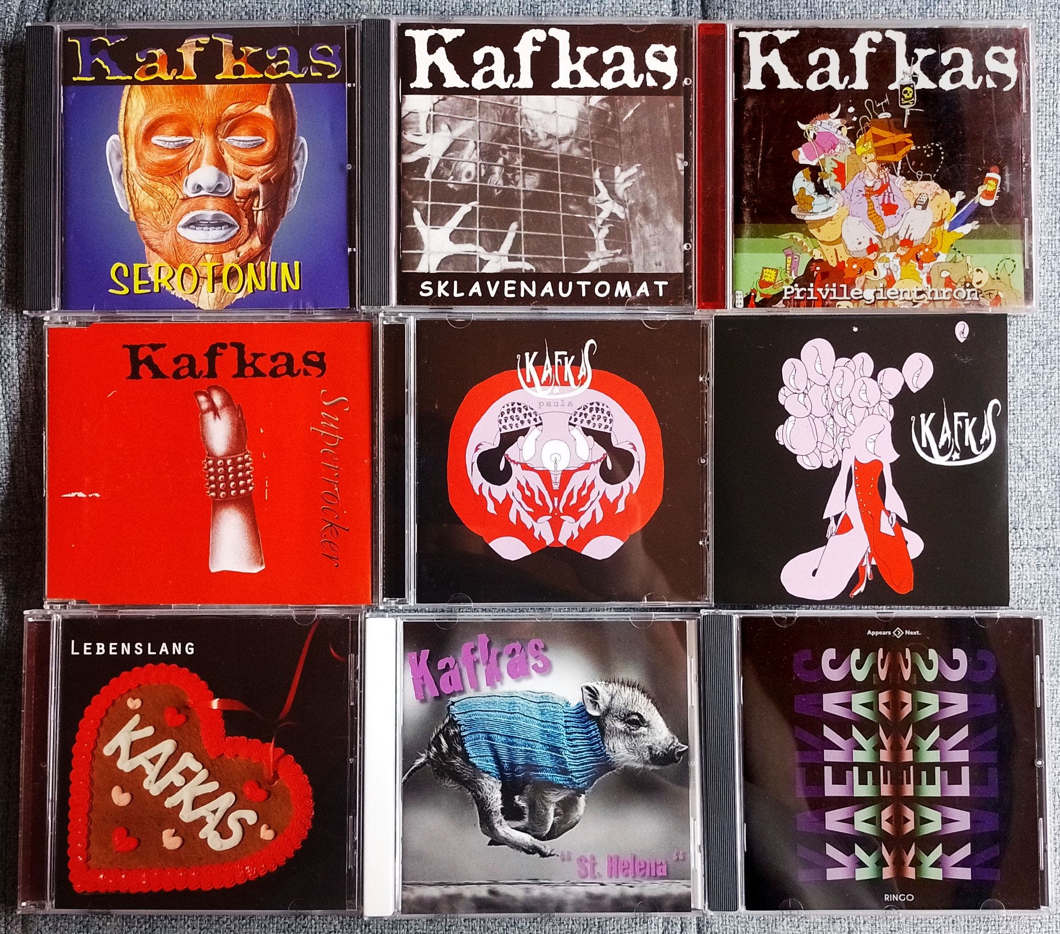 9 CDs by the band Kafkas laid out in a grid: Serotonin, Sklavenautomat, Privilegienthron, Superrocker EP, Paula, Paula promo sampler, Lebenslang EP, St. Helena and Ringo